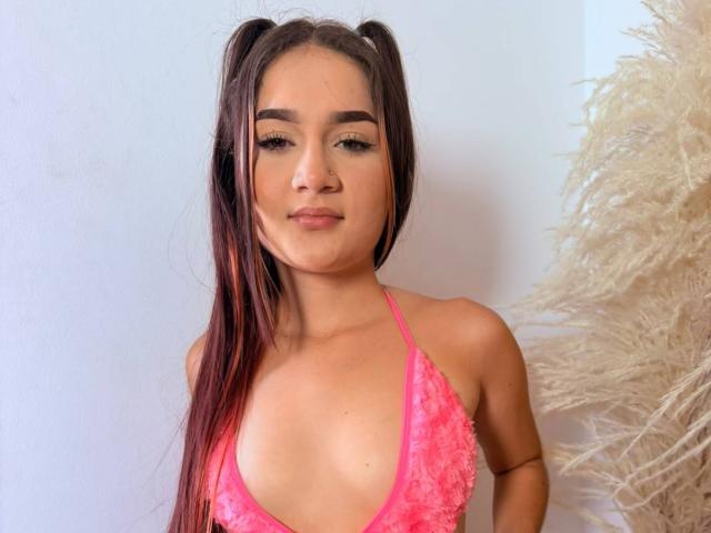 Alisson Miller, con cabello castaño largo en trenzas, vestida con un top rosa de encaje con tirantes, que deja ver sus pequeños pechos, de pie frente a una pared blanca y hierba de pampa esponjosa..
