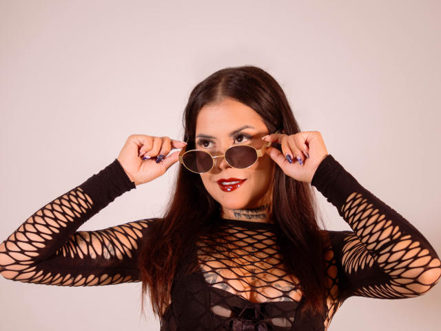 ElectraRoss with long dark hair, wearing a black fishnet top, holding round sunglasses, red lipstick, and blue nail polish, smiling at the camera. Boobs visible through the net.
