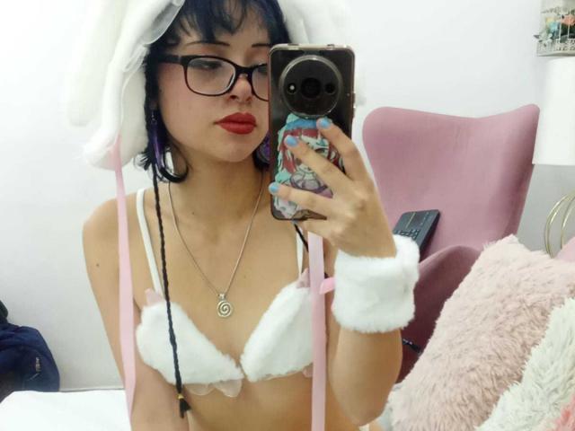 ViolettClay with purple hair, glasses, red lipstick, white furry bra, pink bunny hat, and wrist cuffs, taking a mirror selfie, showing small tits, in a pink chair room.