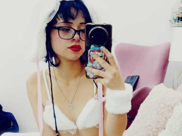 ViolettClay with black hair, glasses, red lipstick, wearing white furry bra and headpiece, holding phone, pink chair, white wall, pink fluffy pillow, visible nipple, ring on chest.