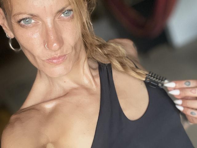 DaneeSoHot hat blaue Augen, einen Nasenring und trägt ein schwarzes Tanktop, das ihre Brüste und ihre muskulösen Arme zeigt. Sie schaut mit einem leichten Grinsen in die Kamera..