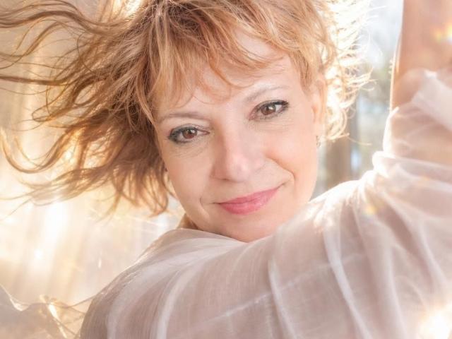 SamantaFocsy, avec ses cheveux bruns clairs et ondulés, emportés par le vent, souriant doucement, portant un haut blanc transparent, un visage illuminé par le soleil, un maquillage discret, des yeux verts et un rouge à lèvres rose..
