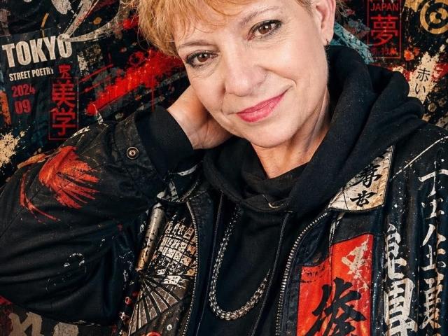 Samanta Focsy, avec ses courts cheveux blonds, souriante, portant une veste en cuir noir avec des graffitis rouges et blancs, et un sweat à capuche noir, a un collier en chaîne et un joli visage..