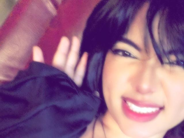 AnnieUwU has dark hair, a cute smile with pink lipstick, and is wearing a black shirt, showing her hand in the blurred background. Her tits are partially visible.