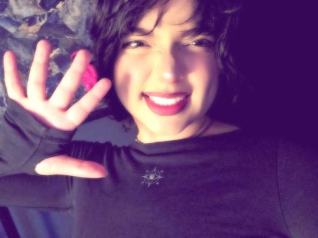 AnnieUwU with short black hair, smiling with red lipstick, holding a pink object in her right hand, wearing a black long-sleeve shirt, looking happy and playful.