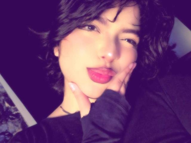 AnnieUwU with short black hair, pink lipstick, and black shirt, tongue out, hand on cheek, smiling. Her nose is slightly upturned. She has a cute, sexy look.
