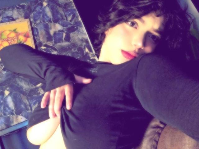 AnnieUwU with dark wavy hair, red lipstick, and black long-sleeve top, covering her small tits with one hand, lying on a couch. Her blue eyes look at the camera.