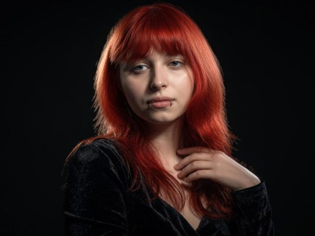 CarolanNiheu, met rood haar, bleke huid en blauwe ogen, draagt een zwarte fluwelen top en houdt haar linkerhand in de buurt van haar nek. Ze kijkt er iets verdrietig uit, tegen een donkere achtergrond..