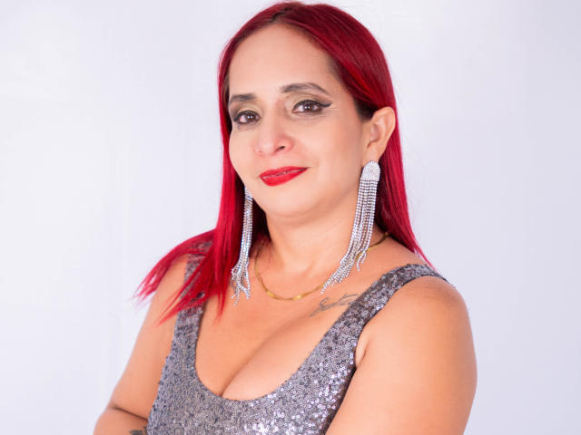 KateMonroe has red hair, red lipstick, and wears a sparkly gray dress with large diamond earrings. Her tits are noticeable in the low-cut dress. She smiles at the camera.