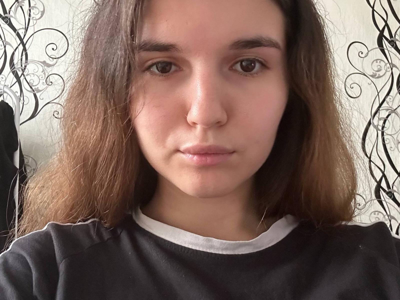 ShellaAleff has a serious face with brown eyes, long brown hair, and a black shirt with white trim. She's looking directly at the camera, with a neutral expression.