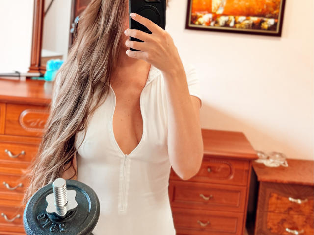 BiancaHotLove si sta scattando un selfie allo specchio, indossando un abito bianco, aderente e con un profondo scollo a V, che mette in mostra i suoi piccoli seni. Ha lunghi capelli castani ondulati e tiene un manubrio davanti a sé..