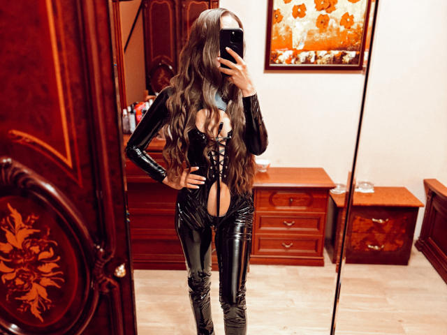 BiancaHotLove con lunghi capelli castani ondulati, un abito in lattice nero con lacci sul petto, che mostra i suoi seni, mentre scatta un selfie allo specchio in una stanza di legno..