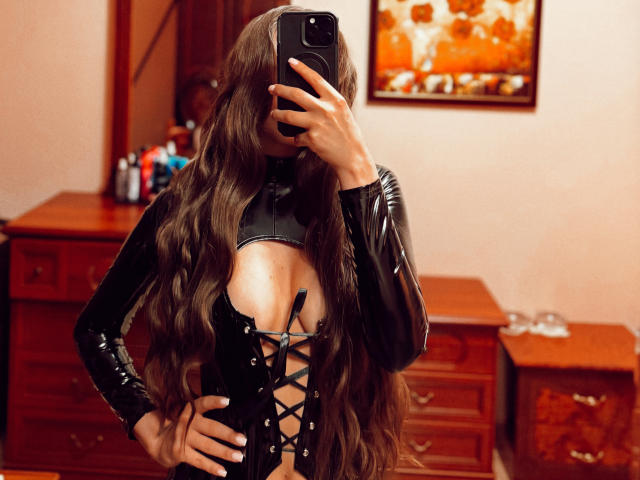 BiancaHotLove with long wavy brown hair, wearing a black leather lace-up top, exposing her big boobs, taking a mirror selfie with a black phone in a wooden room.