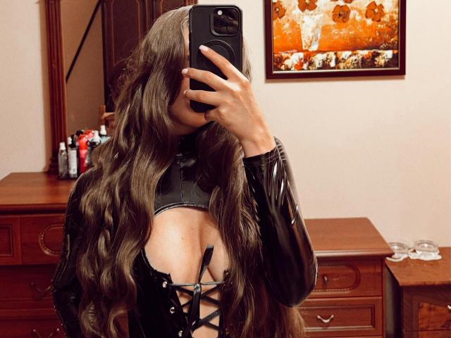 BiancaHotLove, com cabelo castanho ondulado, um corpete de couro preto e seios à mostra, tirando uma selfie no espelho com o celular, em um quarto com móveis de madeira..