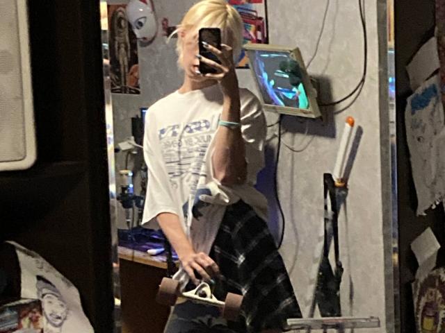 Darline Halasz, con i capelli biondi, una maglietta bianca e pantaloni a quadri, sta scattando un selfie allo specchio con un telefono, tenendo in mano un giocattolo o un telecomando, in una stanza disordinata..