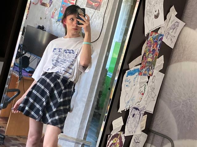 Darline Halasz mit kariertem Rock, weißem T-Shirt mit Aufdruck und schwarzen Haaren, in einem Spiegel-Selfie, wobei ihr Hintern zu sehen ist, sie hält ein Handy, ein Kunstwerk an der Wand, ein Bürostuhl im Hintergrund, sie schaut auf das Handy..