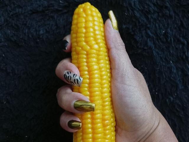 LailaCastillo's hand holds a bright yellow corn on the cob, showing off her gold and black nail polish against a black textured background.