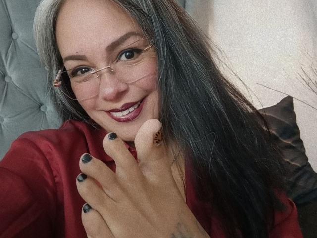 LailaCastillo with long gray-black hair, glasses, red shirt, smiling, holding her foot with black-nail-painted toes, showing a small tattoo on her finger.