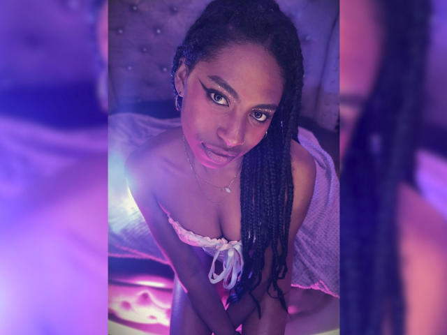 QueenPerlaa mit dunkler Haut, langen schwarzen Zöpfen und lila Eyeliner, die mit einem leichten Lächeln nach oben schaut, trägt ein weißes Top mit einem Schulterfrei-Schnitt, das kleine Brüste zeigt, in einem violett beleuchteten Raum..