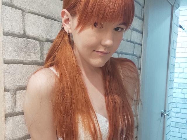 SheritaChieng, con cabello rojo, blusa blanca de encaje, pechos pequeños, piel clara, flequillo, perforación en la oreja, ligera sonrisa, de pie cerca de una pared de ladrillos blancos, mirando a la cámara..