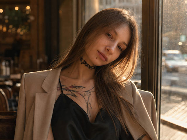 JaPorada with long brown hair, wearing a black satin top and beige blazer, choker, tattoo on chest, looking down, soft sunlight, window, urban street outside, casual, sexy.