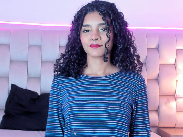 RayliCurly has curly black hair, brown eyes, and a nose ring. She's wearing a blue striped shirt, sitting in front of a padded, light-colored headboard.