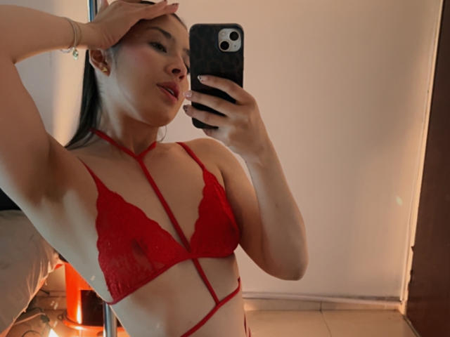 EmmaLombardi taking a mirror selfie, wearing a red lace bra with strappy details, her hand on her head, showing her tits and a cute smile, holding a black phone.