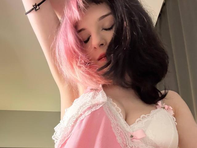 ChangCaddle with pink and black hair, eyes closed, wearing pink lace lingerie, arm raised, showing small tits, pink bow, white lace trim, black bracelet, soft light.