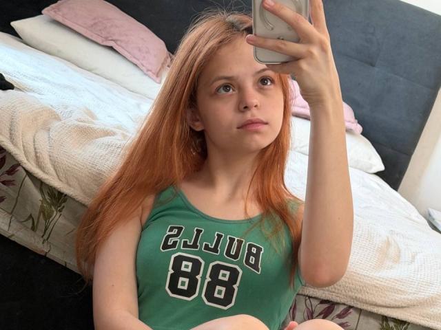 Sadie Yankovitz, avec de longs cheveux roux, un débardeur vert avec le chiffre "88" en noir, tenant un téléphone, prenant un selfie devant un miroir, regardant vers le haut, un lit avec un oreiller rose en arrière-plan..
