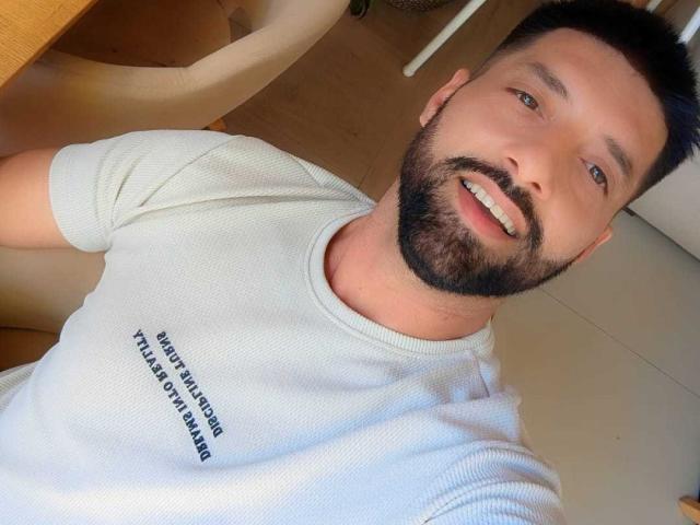 TommyDum with a beard, short black hair, smiling, white ribbed t-shirt, text on shirt, sitting, wooden table, light wood floor, casual selfie, blue eyes, attractive.