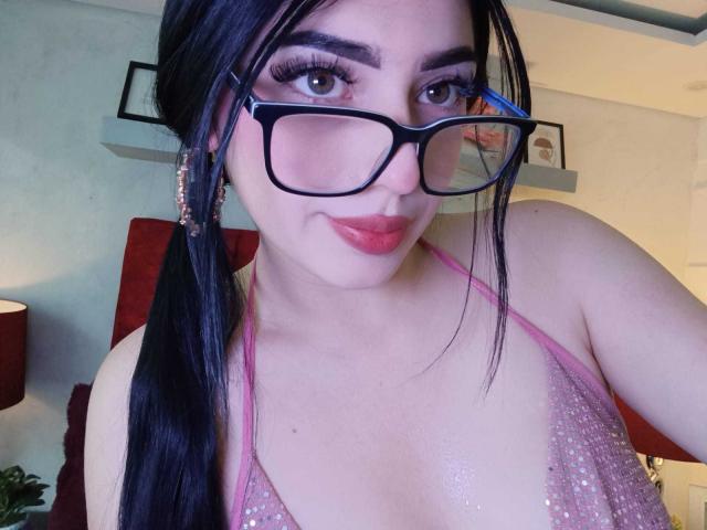 Isabella Rossi met een zwarte paardenstaart, een bril, een roze glinsterende haltertop, die haar borsten laat zien, roze lippenstift, eyeliner en oorbellen, lachend naar de camera..