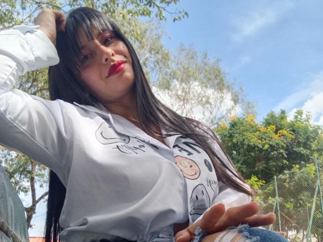 MadissonFontaine with long black hair, red lipstick, white shirt with smiley face print, hand in hair, blue jeans, and ring, sitting outdoors, clear sky, trees, and fence.