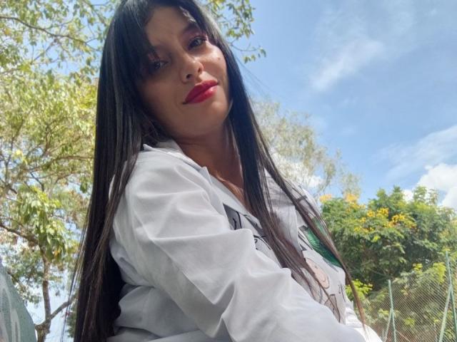 MadissonFontaine with long black hair, red lipstick, and white shirt, looking down with a slight smile, outdoors, under a blue sky with scattered clouds.