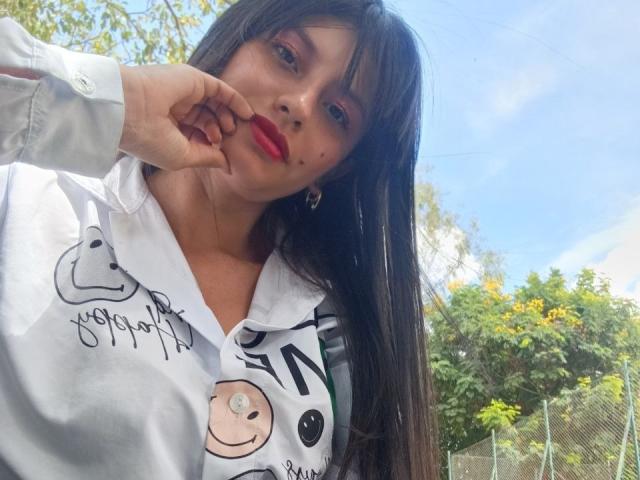 MadissonFontaine with long black hair, red lipstick, white shirt with cute doodles, and a ringed nose, touching her lip with her finger, outside on a sunny day.