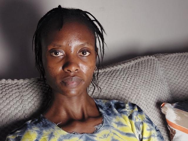 CandyFlossy, con piel oscura, rostro brillante y nariz pequeña, lleva una camiseta con estampado tie-dye, tiene trenzas cortas y negras, y está sentada en un sofá gris con textura..