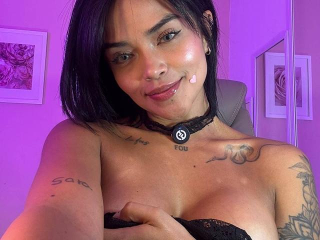 MajoJones con cabello negro, un pequeño corazón rosa en la mejilla, un collar negro con el logo de Spotify, tatuajes en el pecho y el brazo, sosteniendo senos cubiertos de encaje negro, sonriendo a la cámara..