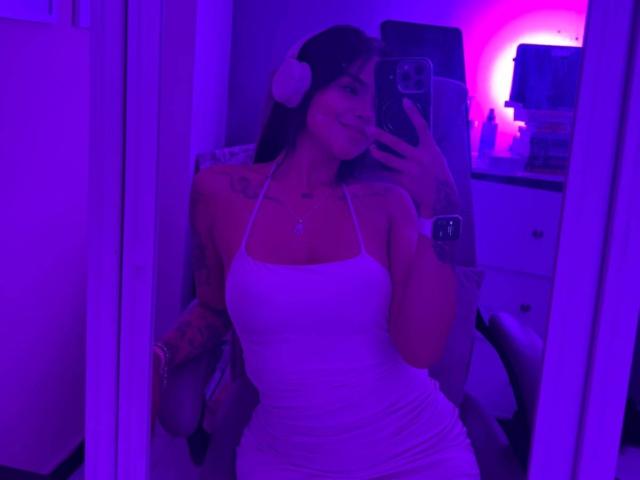 MajoJones in a white halter dress, showing her tits, with tattoos on her arms, taking a mirror selfie, purple neon lighting, smiling, phone in hand.