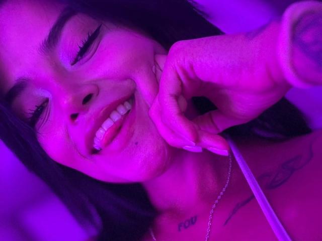 MajoJones con una sonrisa, iluminación púrpura, mano tocándose la barbilla, tatuajes visibles, cabello largo y oscuro, delineador de ojos, blusa blanca, collar, dientes y una nariz linda. Sus glúteos no son visibles..