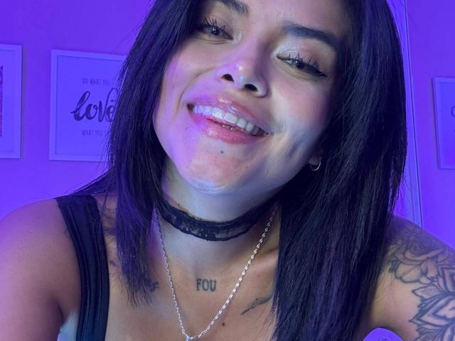 MajoJones with black hair, smiling, glossy lips, black lace choker, tattoos, "FOO" on chest, purple lighting, necklace, visible cleavage, earrings, tattoos on shoulder, and arm.