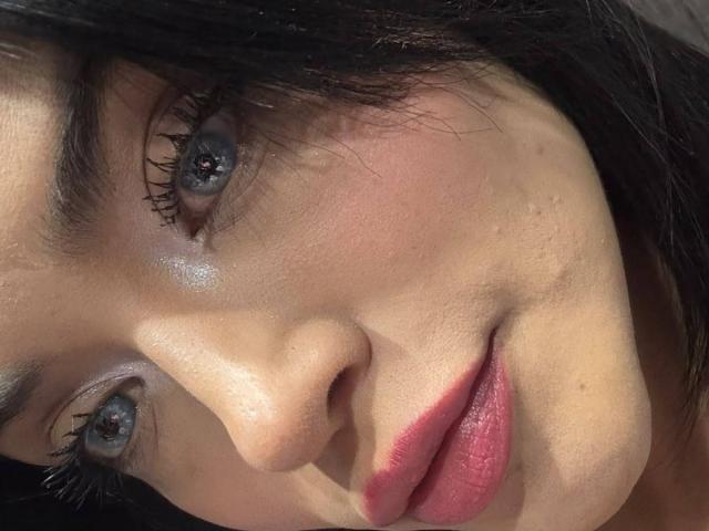 MajoJones's face with blue eyes, pink lips, slight blush, and dark eyelashes, showing a soft smile. Her skin is smooth with small water droplets. She's looking at the camera.