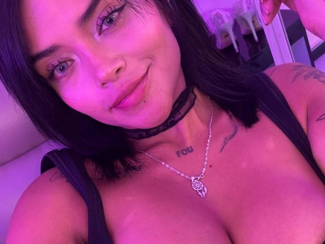MajoJones with black hair, purple lighting, black choker, necklace, tattoos, and a smile, showing large tits and a cute nose ring, in a selfie.