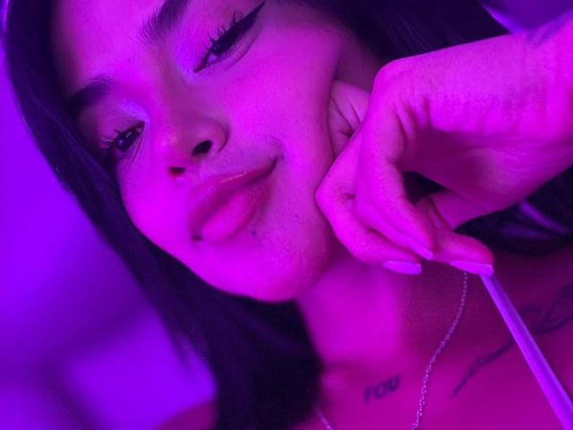 MajoJones with pink-purple lighting, holding phone to ear, slightly pouty lips, dark hair, tattoo on chest, eyeliner, and a necklace, showing a relaxed, confident expression.
