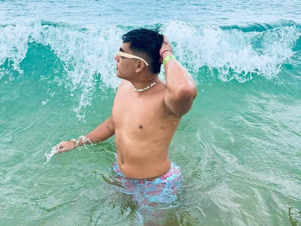 Andy Carrintong, torse nu, portant un maillot de bain coloré, un bandeau jaune et un collier, se tient dans une eau de mer verte, levant la main droite, avec une petite vague derrière lui..
