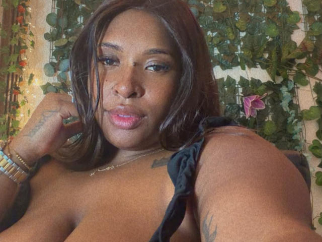 NatyLm, com cabelo castanho, pele escura e batom rosa, exibindo seus seios em uma blusa preta, com tatuagens visíveis, usando pulseiras e olhando para a câmera com um leve sorriso..