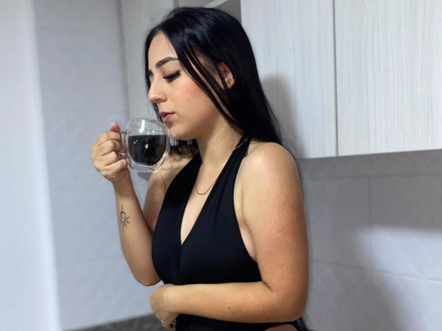StefanyDelly with black hair, wearing a black halter top, holding a glass of dark coffee, tattoo on left wrist, standing in a white-walled room, looking down.