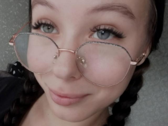 FayStorbeck, com óculos redondos cor-de-rosa, pele clara, olhos azuis, cílios longos e tranças escuras, sorrindo levemente, mostrando os lábios cor-de-rosa, e seu rosto é o foco principal..