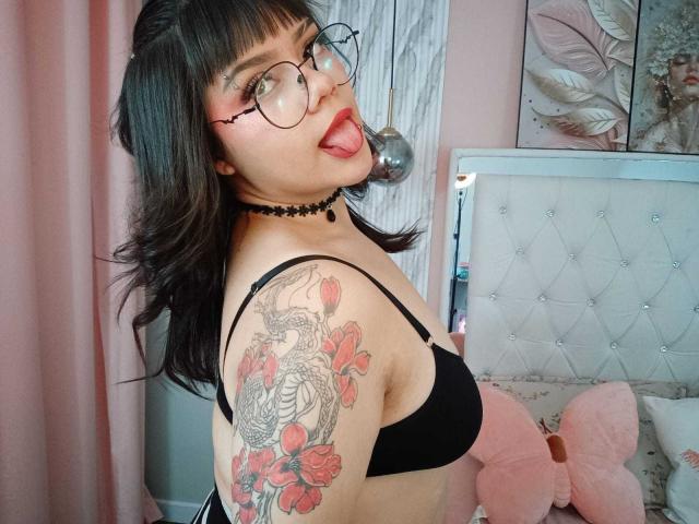 AnneStonee, avec un soutien-gorge noir, la langue tirée, un collier, des lunettes, et un tatouage de dragon sur son bras, exhibant ses seins dans une chambre rose..
