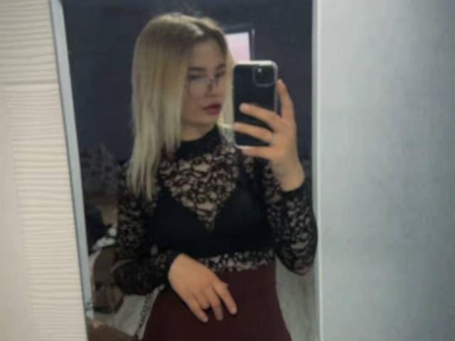 AdrieneDegennaro with blonde hair, red lipstick, black lace top, and maroon high-waisted skirt, holding phone, mirror selfie, showing her tits, in a room.
