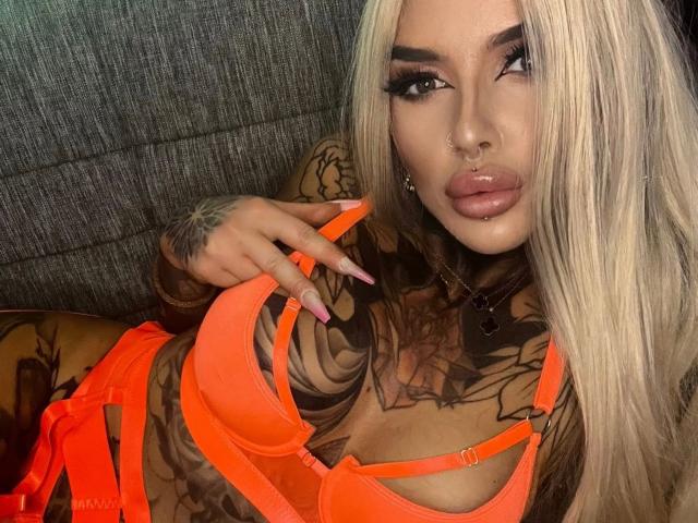 Lexxis-Blck, uma mulher atraente com cabelos loiros longos, piercing no nariz e no lábio, vestindo lingerie laranja neon, exibindo tatuagens e unhas rosa, olhando para cima com lábios cheios e brilhantes..