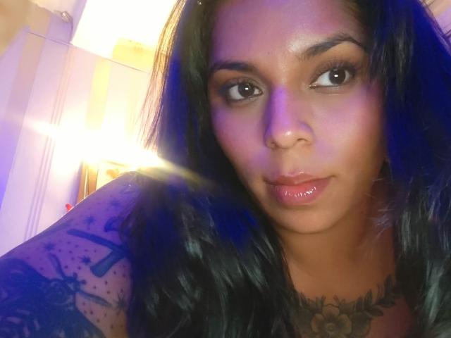 Chili has dark, wavy hair, brown eyes, glossy pink lips, and a tattoo on her right arm showing a gun. She's looking at the camera with a subtle smile.
